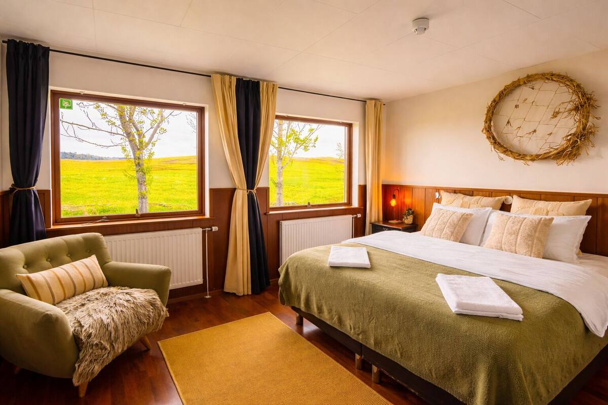 Une chambre d’hôtel avec un lit, une chaise, une fenêtre avec des rideaux, un radiateur et une horloge murale.