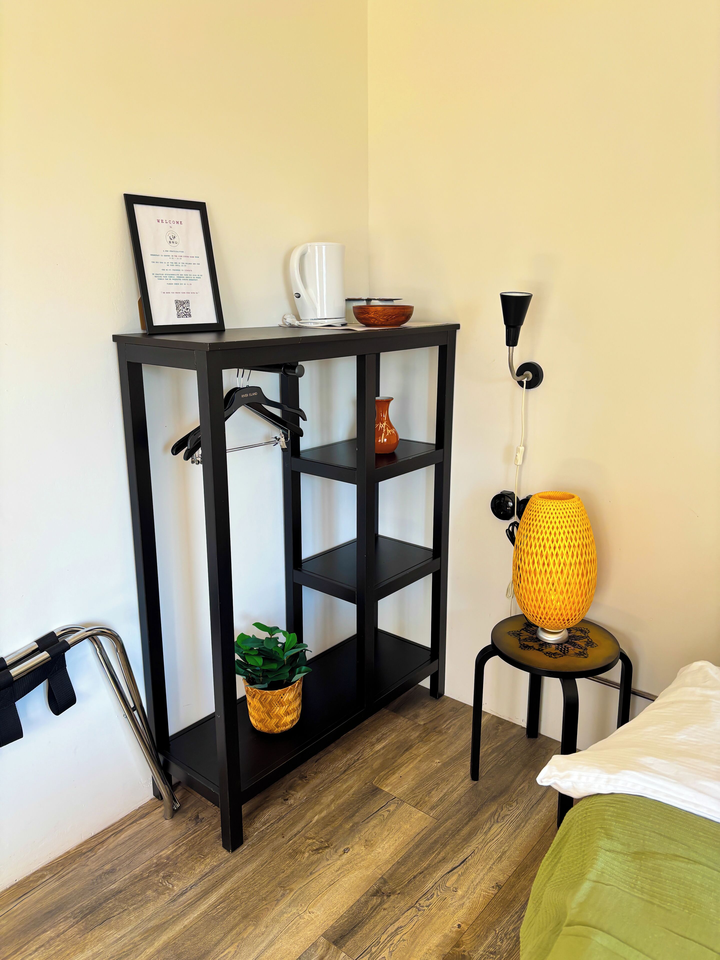 Una habitación pequeña y ordenada con una estantería metálica negra que contiene un letrero de bienvenida en un marco, una cafetera blanca y un cuenco marrón. Una lámpara amarilla está sobre un taburete pequeño y una planta en maceta sobre la estantería.