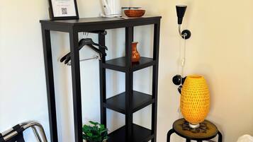 Una habitación pequeña y ordenada con una estantería metálica negra que contiene un letrero de bienvenida en un marco, una cafetera blanca y un cuenco marrón. Una lámpara amarilla está sobre un taburete pequeño y una planta en maceta sobre la estantería.