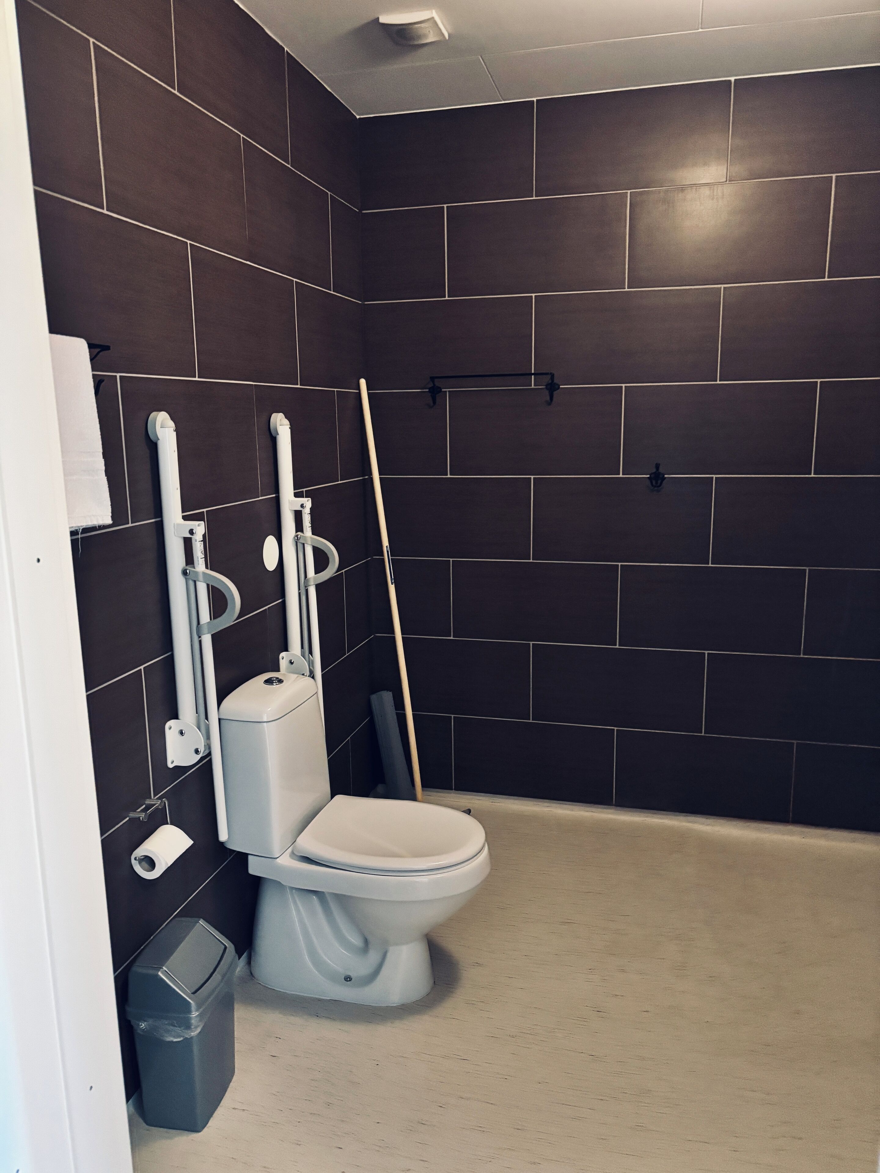Un baño moderno con paredes de azulejos oscuros, un inodoro blanco, un cubo de basura y una escoba apoyada en la pared.
