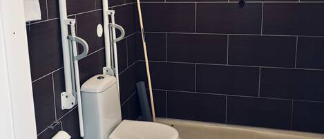 Un baño moderno con paredes de azulejos oscuros, un inodoro blanco, un cubo de basura y una escoba apoyada en la pared.