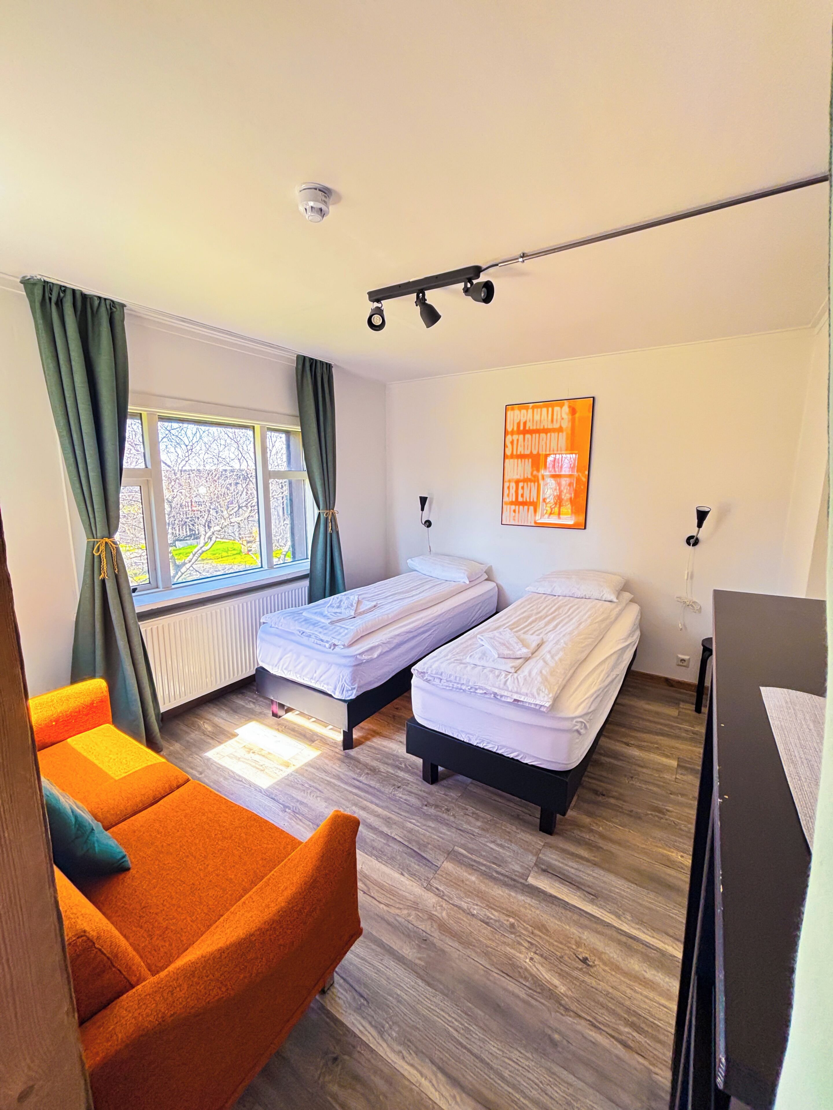 A room with two single beds, a sofa, a radiator, a window with curtains, a ceiling light, and a wall-mounted artwork.