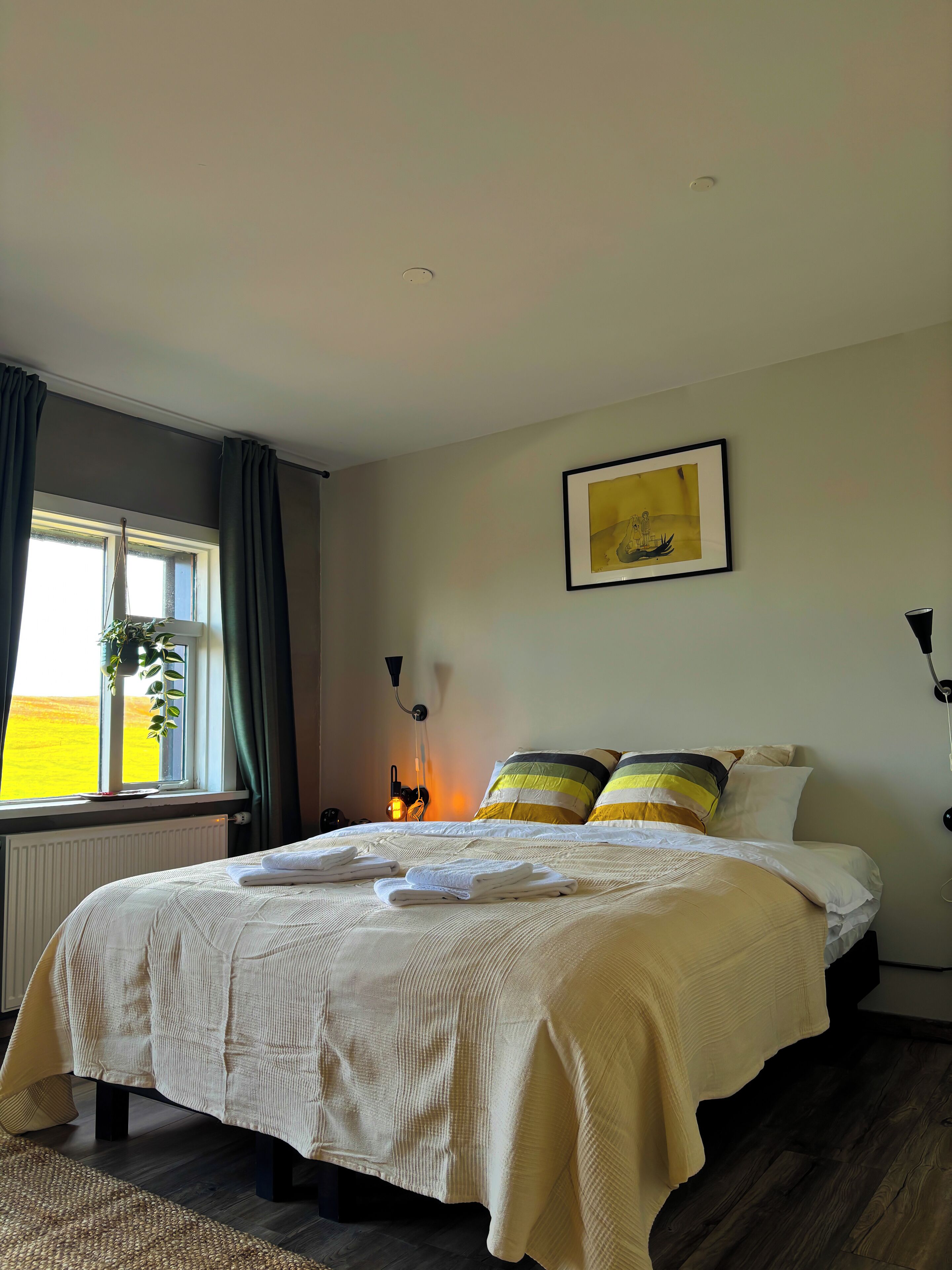 A neatly made bed with a white comforter and pillows, a bedside lamp, a framed picture on the wall, and a window with curtains.