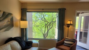 A room with a beige leather sofa, a brown armchair, a large window with a view of trees, and a lamp on the right. - Perfect 2nd floor condo with mtn & golf  views! Charm galore awaits you! (Tucson)