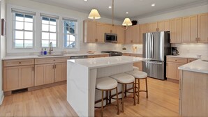 A modern kitchen with wooden cabinets, stainless steel appliances, a central island with bar stools, and pendant lighting. - Dockside Hideaway: Enjoy Privacy and Scenic Views (Wrightsville Beach)