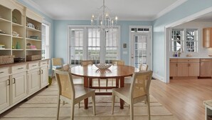 A dining room with a round table, six chairs, a chandelier, and a view through large windows. - Dockside Hideaway: Enjoy Privacy and Scenic Views (Wrightsville Beach)