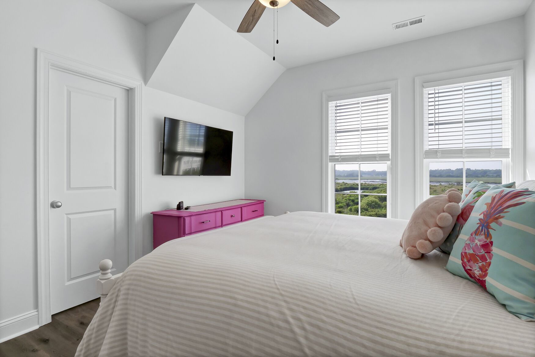 A bedroom with a bed, a pink dresser, a flat-screen TV, and a view of the outdoors through the windows.