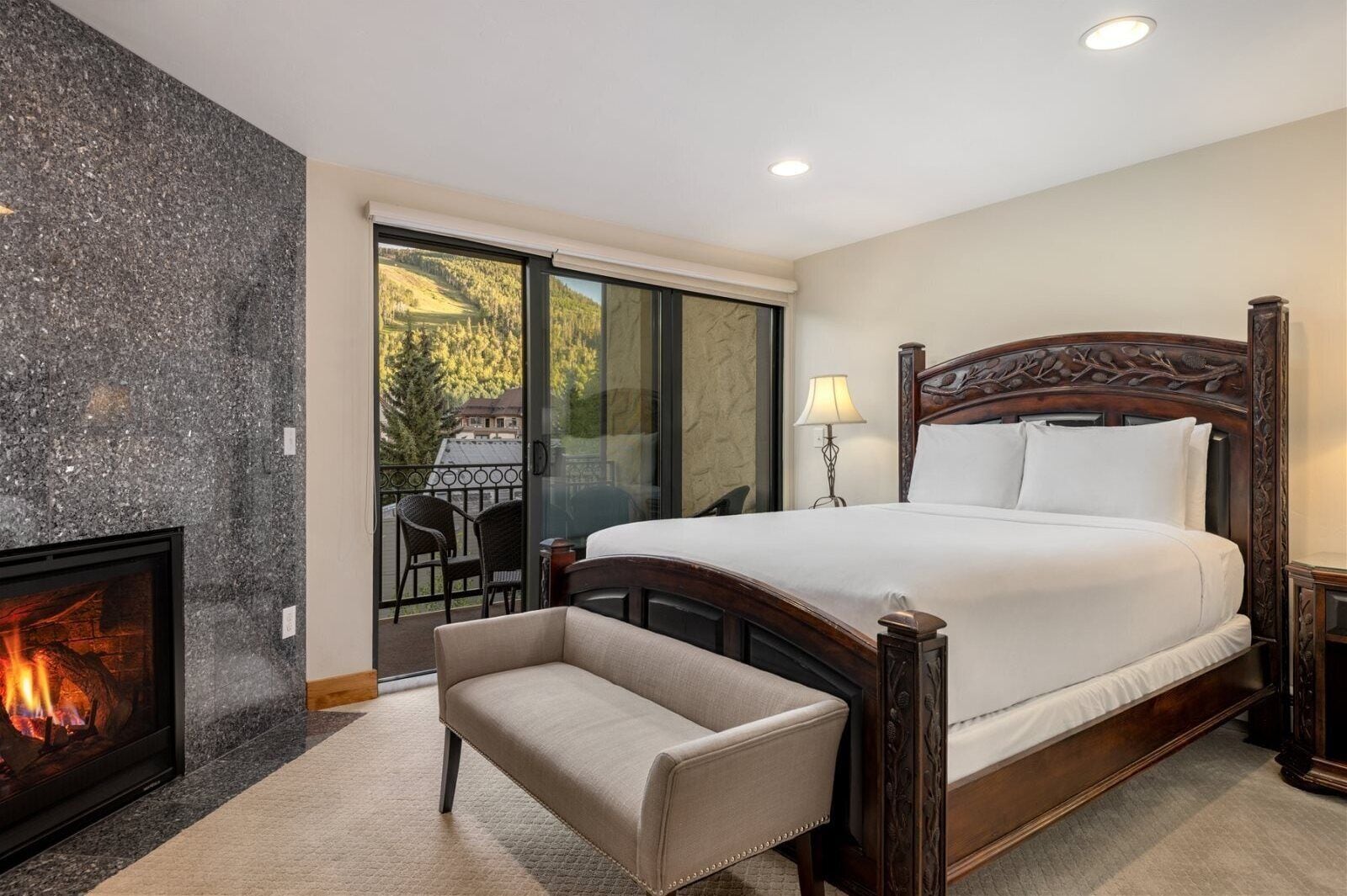 A bedroom with a large bed, a fireplace, a balcony with a view of trees, and a chair.