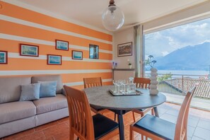 Un coin repas avec une table ronde, des chaises et un canapé. Des tableaux encadrés ornent le mur, et la fenêtre offre une vue sur une montagne et un lac.