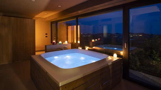 A modern bathroom with a large bathtub, a vanity, and a window offering a city view at dusk. - Relais Casa Clara (Monteu Roero)