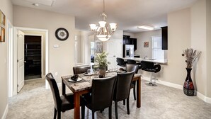 A dining area with a chandelier, a dining table set for four, and a kitchen in the background. - 4329ac - Solterra Gated Resort 5 Bedroom Home by RedAwning (Davenport)
