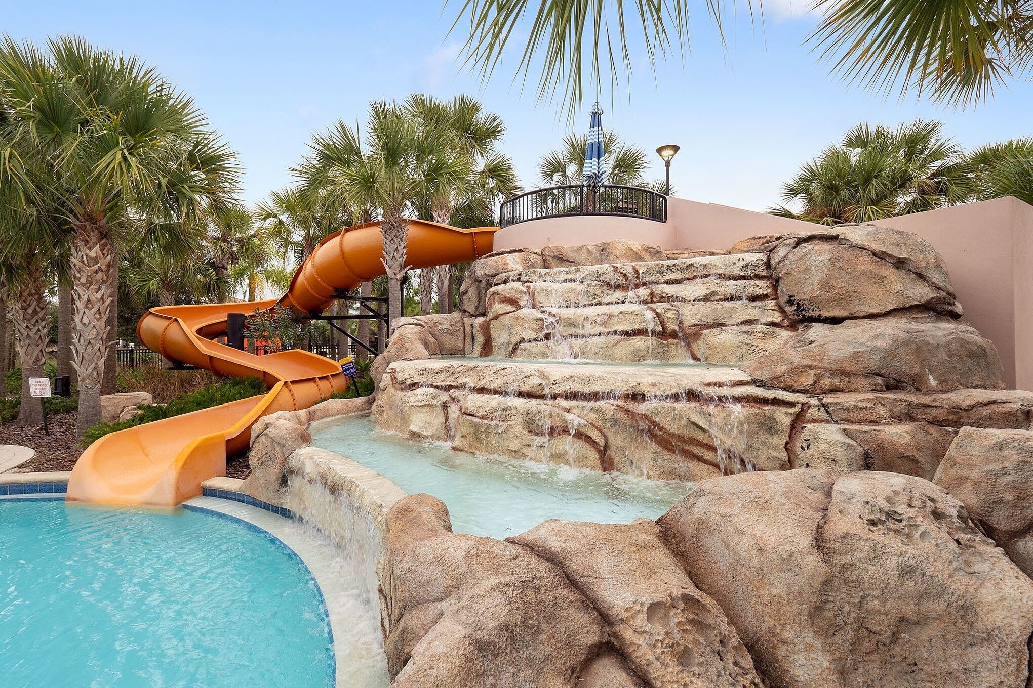 A water slide leading into a pool, surrounded by palm trees and a rocky landscape.