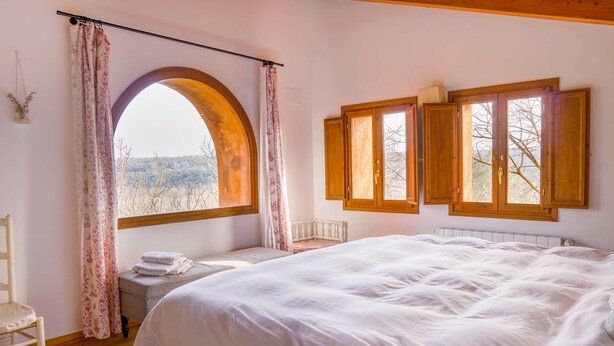 Un dormitorio con una cama grande, un banco de madera, una ventana con cortinas y vistas al exterior.