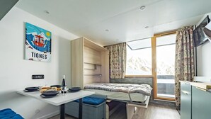 A compact room with a bed, a small table with a bottle and glasses, a blue bench, and a window with curtains. - 100m from resort center, Ground floor, balcony, tv, ski locker, 21m², Tignes (Tignes)