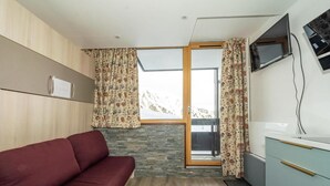 A compact room with a red sofa, a small kitchenette, a TV mounted on the wall, and a window with patterned curtains. - 100m from resort center, Ground floor, balcony, tv, ski locker, 21m², Tignes (Tignes)