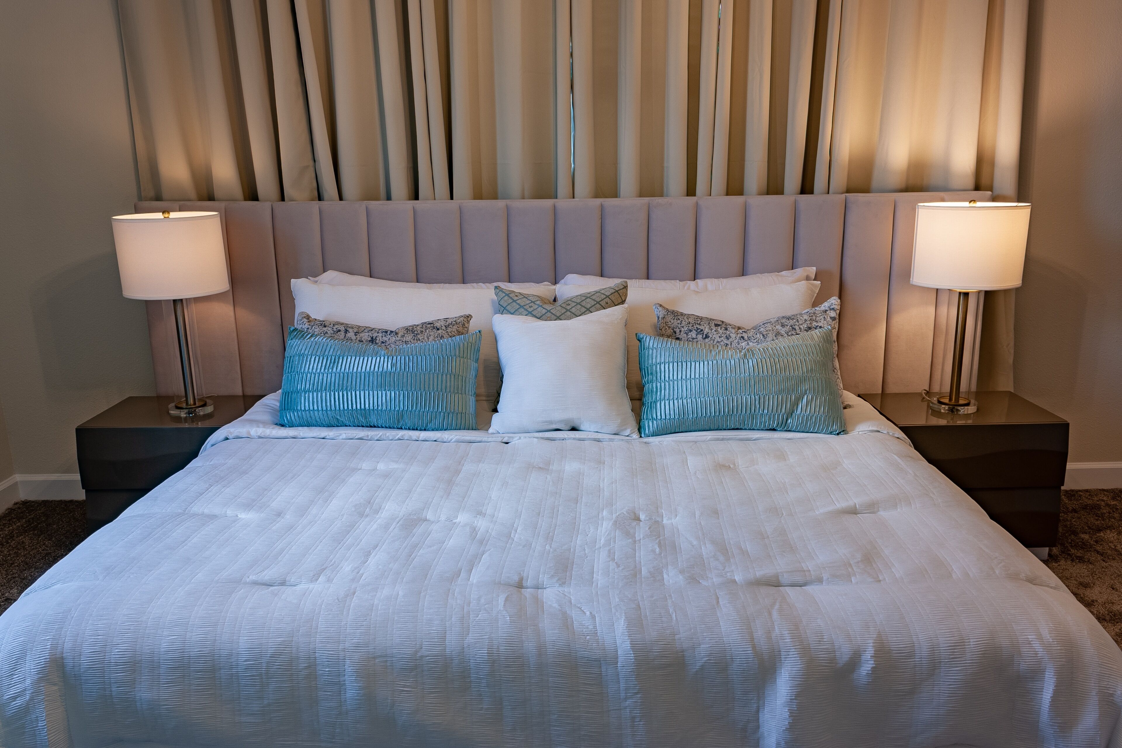 A neatly made bed with a striped white comforter, two matching white pillows, and three decorative pillows with blue and white patterns. There are two bedside tables with lamps on either side of the bed.
