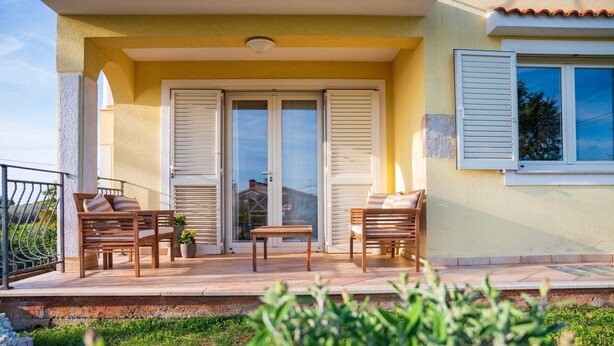 Un balcón con dos sillas de madera, una pequeña mesa, un ventanal corredero y una ventana con contraventanas.