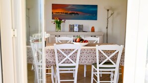 Un comedor con una mesa preparada para cuatro personas, sillas blancas, un mantel con estampado floral y un frutero.