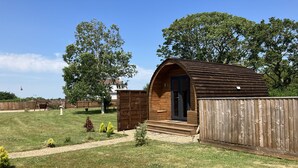 Eine kleine Holzhütte mit einer dunklen Tür, umgeben von einem Holzzaun, in einem grünen Hof mit Bäumen und blauem Himmel.