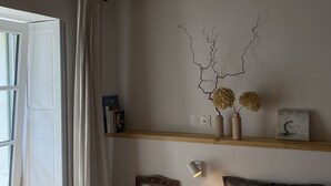 A bedroom with exposed wooden beams, a bed with a white pillow, a wooden chair, a bedside table with books and a vase, and a wall-mounted light.