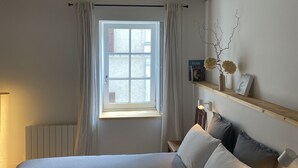 A bedroom with a large bed, a window with curtains, a radiator, and a shelf with decorative items.