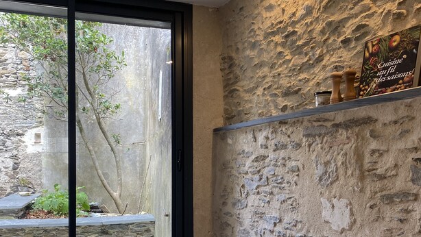 A room with a stone wall, a window with a view of greenery, a small table, and a shelf with items.