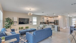 A modern living room with a large TV, blue sofas, and a kitchen in the background. - 1233chd - The Retreat At Championsgate 9 Bedroom Home by RedAwning (Davenport)
