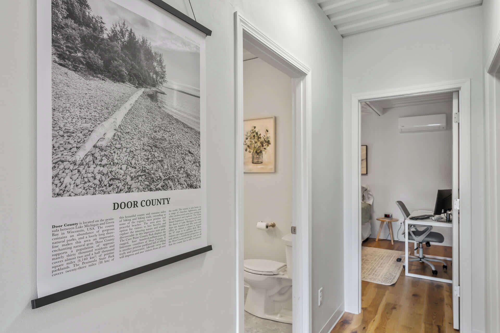 En gang med et indrammet kort over Door County, et indrammet blomsterbillede og et glimt af et badeværelse med et toilet og et skrivebord.