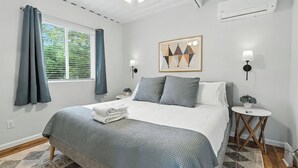 Ein Schlafzimmer mit Bett, Deckenventilator, Klimaanlage, einem Fenster mit Vorhängen, einer Wandlampe und einem kleinen Tisch mit Pflanze.