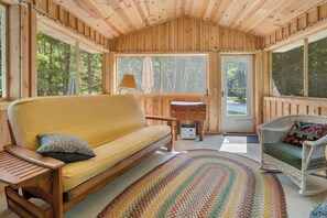 A wooden cabin interior with a sofa, a wicker chair, a small table, and a large window.