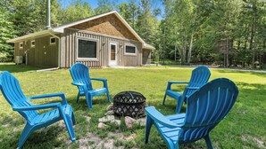 Une cabane avec une façade en bois, une cheminée et un foyer entouré de quatre chaises Adirondack bleues.