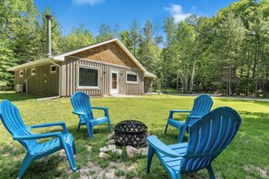 A cabin with a wooden exterior, a chimney, and a fire pit surrounded by four blue Adirondack chairs.