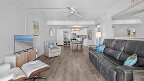 A spacious living room with a grey leather sofa, a white armchair, and a wooden bench. A dining area with a table and chairs is visible in the background. - 🌊Magnificent Ocean Views-Pool-Castle Reef 429🌊 (New Smyrna Beach)