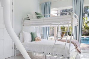 A bunk bed with a white frame, a white mattress, and a beige blanket. There are pillows in various colors.