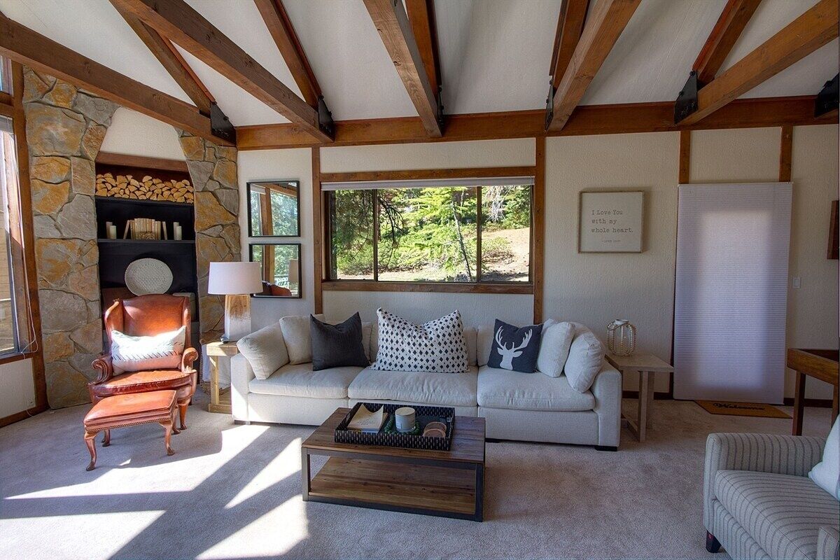 A cozy living room with a stone fireplace, a wooden ceiling, and a large window offering a view of greenery.