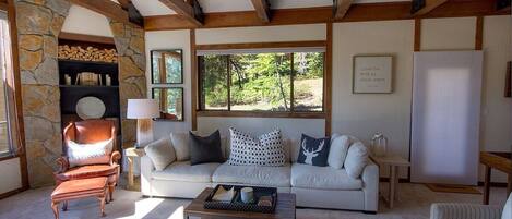 A cozy living room with a stone fireplace, a wooden ceiling, and a large window offering a view of greenery.