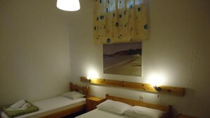 A room with two single beds, wooden headboards, a nightstand with a lamp, a small table with a towel and a book, and a framed picture on the wall.