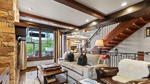 A spacious living room with a stone wall, a wooden staircase, a leather sofa, and a large window offering a view of the outdoors. - 1138 Skiers Heaven! Luxury Home Close To Park City Mountain! Two Hot Tubs & Gorgeous Outdoor Space! 6 Bedroom Home by RedAwning (Park City)