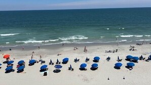 Ein Strand mit Reihen blauer Sonnenschirme und Liegestühle, und einige Personen flanieren am Ufer entlang.