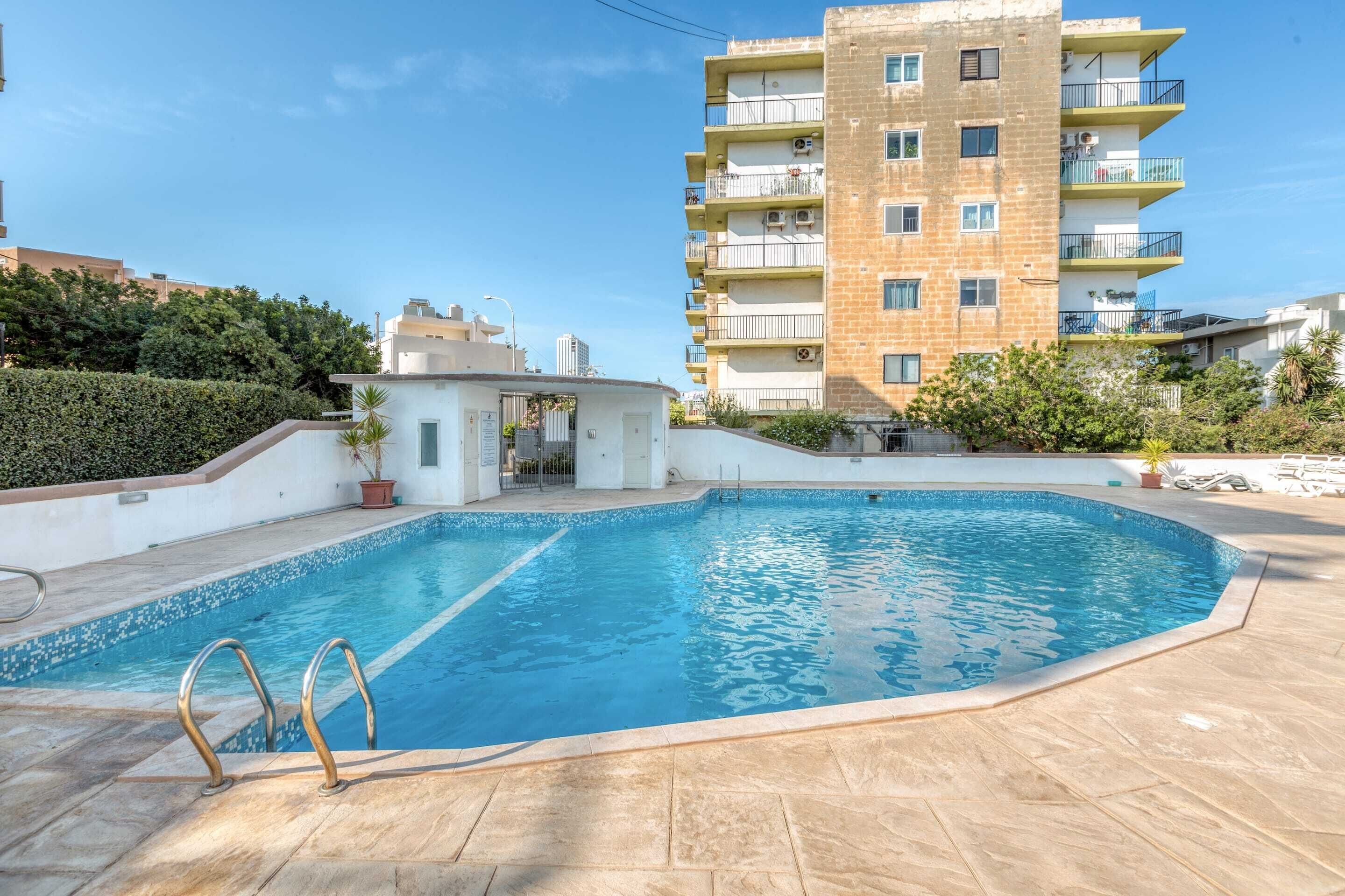 A swimming pool with a tiled deck, surrounded by a low wall and a small building.