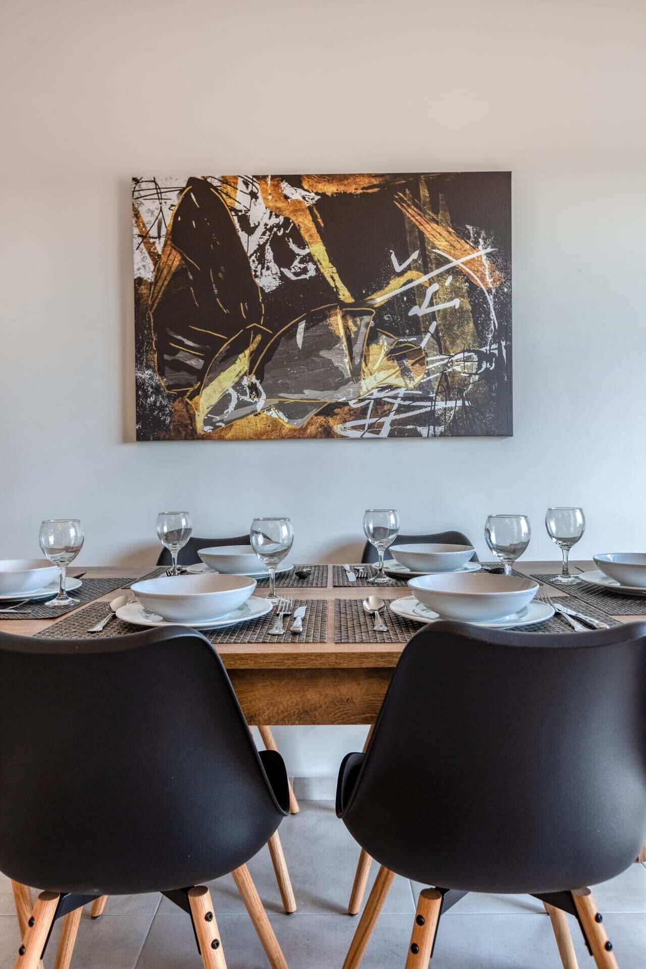 A dining table set with white plates, clear wine glasses, and black chairs with wooden legs.