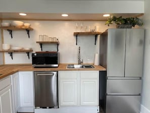 Una cocina moderna con armarios blancos, electrodomésticos de acero inoxidable, encimeras de madera y estantes colgantes con vajilla.
