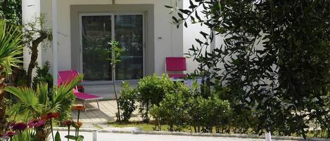 Un edificio moderno con fachada blanca, puerta de cristal y un pequeño patio con sillas rosadas.
