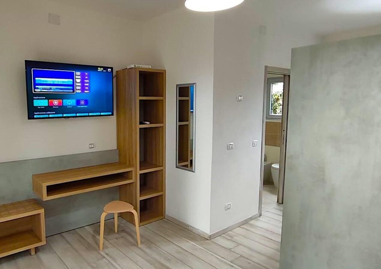 Quarto com televisão fixada na parede, estantes de madeira, um banquinho pequeno e uma porta que dá para outro cômodo.