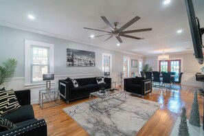 A spacious living room with a ceiling fan, a large window, a black sofa, a glass coffee table, and a dining area with chairs and a table.