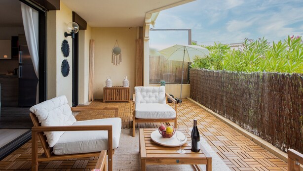 A balcony with a wooden floor, white cushioned chairs, a table with a fruit bowl and a bottle, and a large umbrella. - Happyfew La terrasse de Flora (Nice)