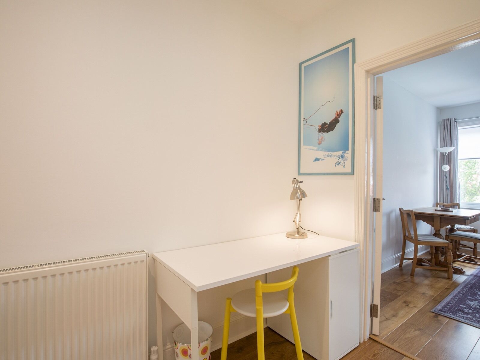 A room with a white desk, a yellow chair, a radiator, a small trash bin, and a door leading to another room.