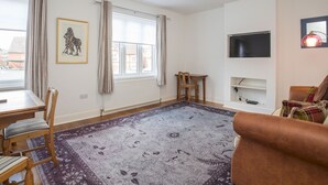 Ein Wohnzimmer mit einem Holztisch, Stühlen, einem gemusterten Teppich, einem braunen Sofa, einem Fernseher und einem gerahmten Bild an der Wand.
