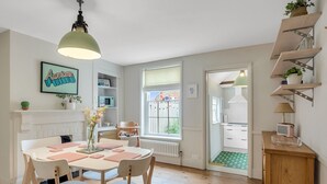 Uma sala de jantar luminosa e bem iluminada, com uma mesa redonda, cadeiras e um lustre pendurado. É visível uma janela com vista para o exterior, e existem prateleiras com diversos objetos na parede.