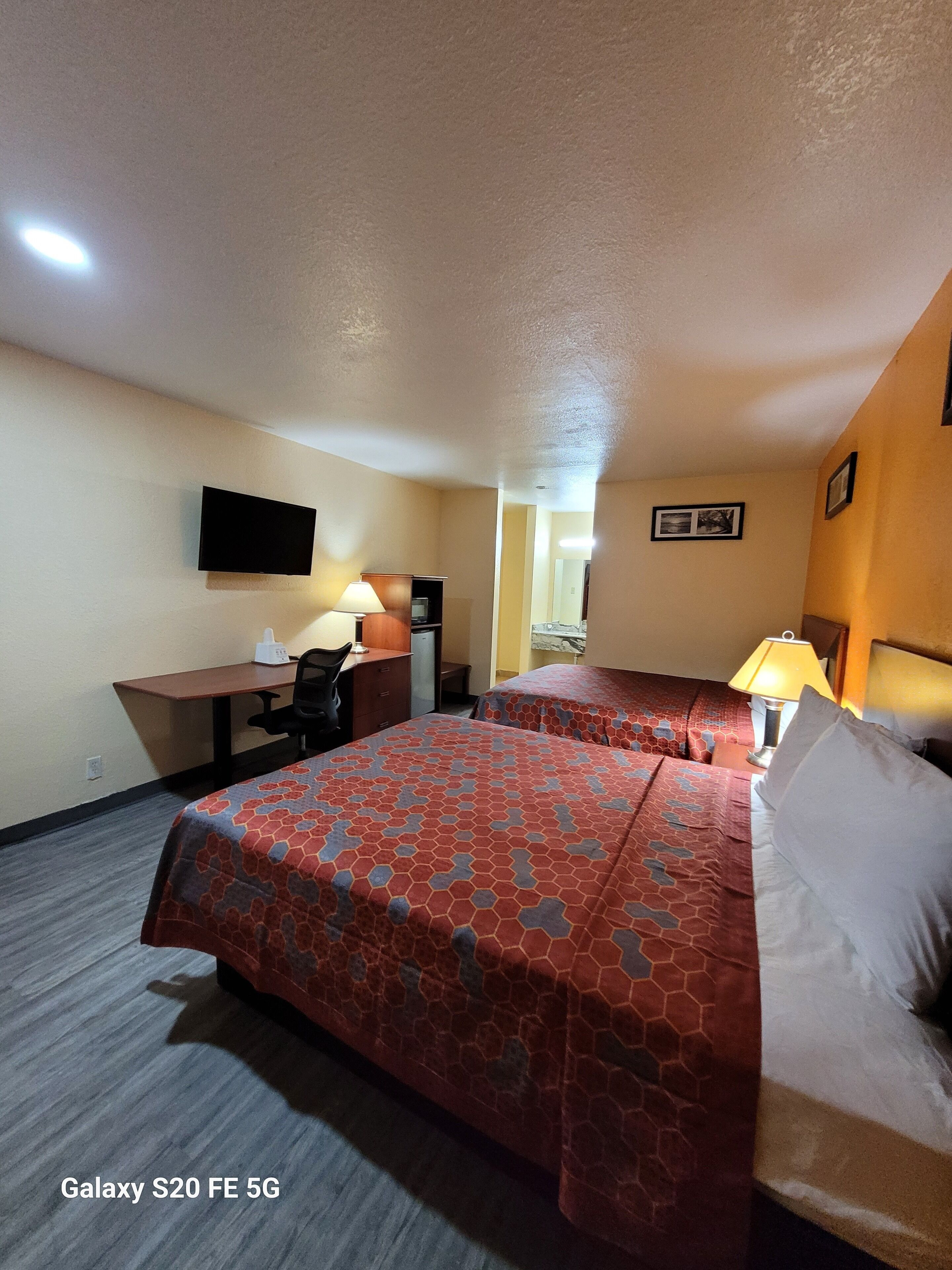 A hotel room with two beds, a desk, a chair, a TV, and a lamp.